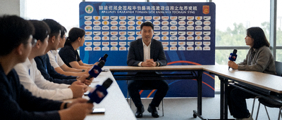 足球新闻发布会现场，主教练坐在桌前面对众多记者回答问题，背景板上印有赛事和赞助商的标志
